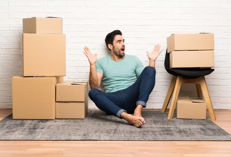 Handsome young man moving in new home among boxes with surprise facial expression