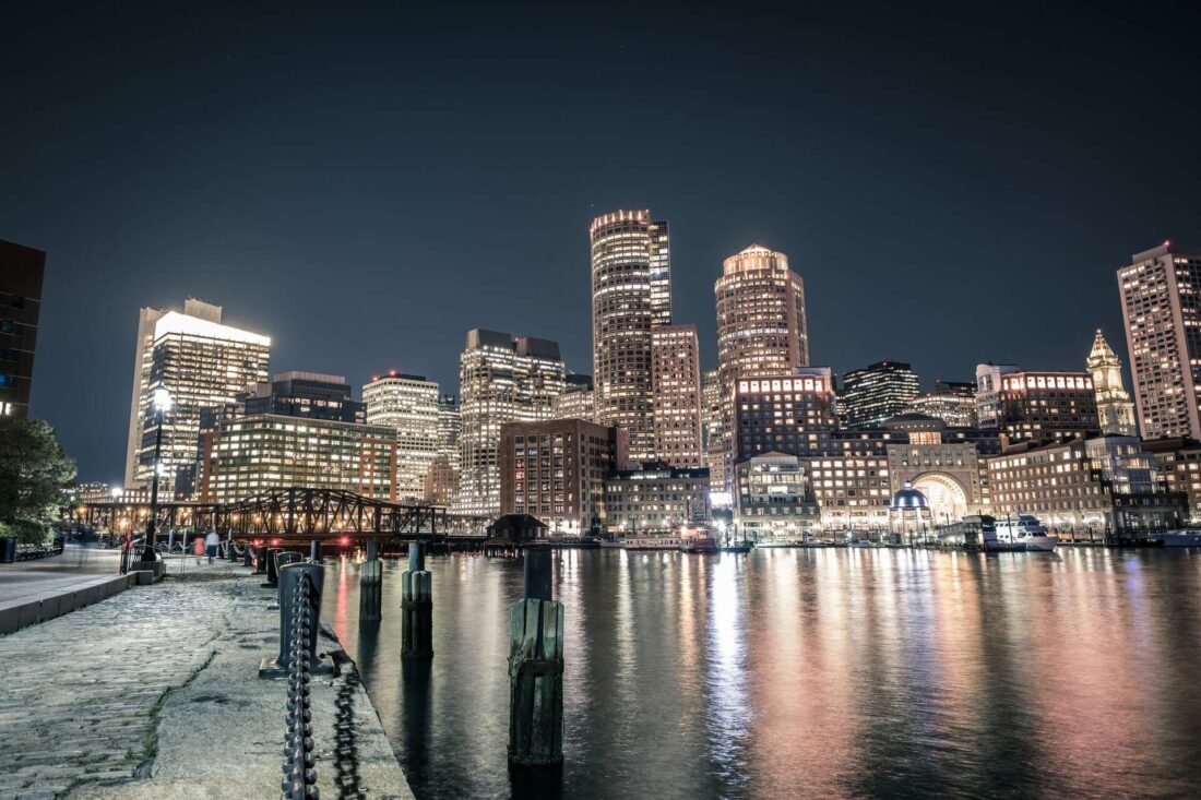 Illuminated Boston skyline viewed from the river shore at night, where the city lights reflect in the calm waters. The darkened sky serves as a backdrop to the glittering cityscape, creating a captivating and enchanting scene that showcases the beauty of Boston's nighttime ambiance along the river