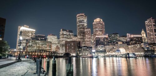 Illuminated Boston skyline viewed from the river shore at night, where the city lights reflect in the calm waters. The darkened sky serves as a backdrop to the glittering cityscape, creating a captivating and enchanting scene that showcases the beauty of Boston's nighttime ambiance along the river