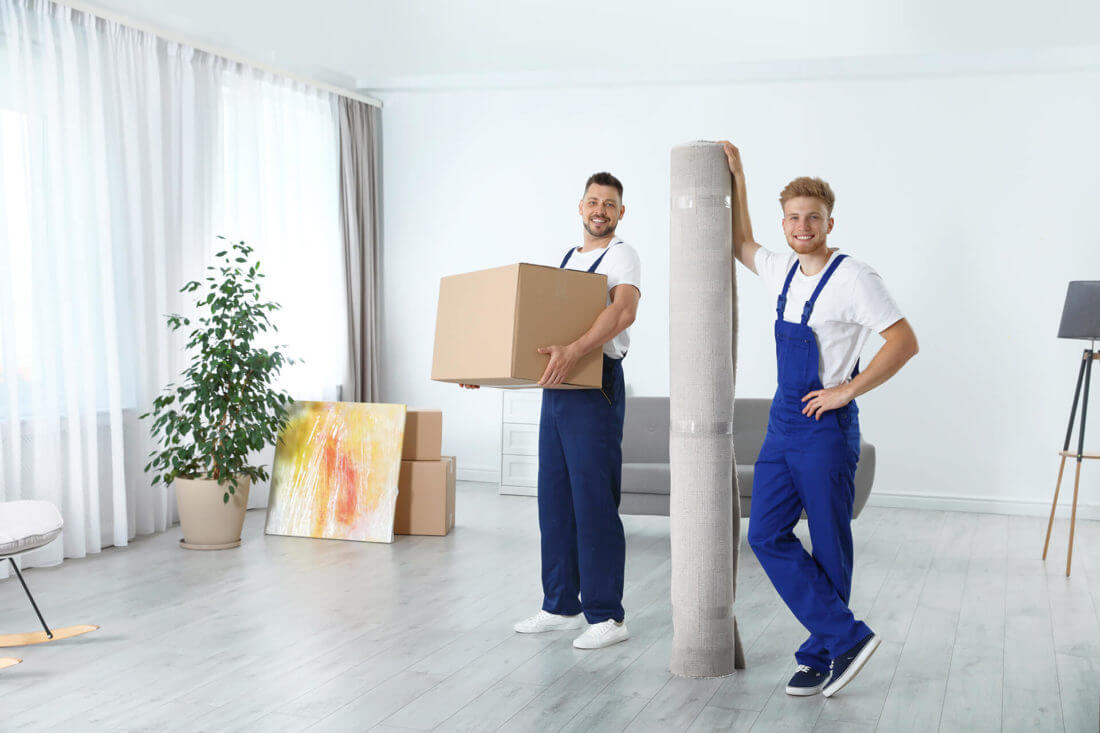 Professional movers in uniforms are standing in the living room, holding boxes and rugs 