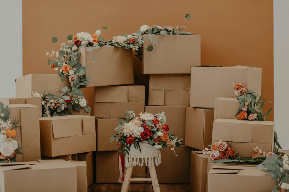 Boxes juxtaposed with vibrant flowers, a scene of transition and renewal. The boxes represent the practicalities of moving, while the flowers symbolize the fresh beginnings and a touch of beauty amidst the process of change.