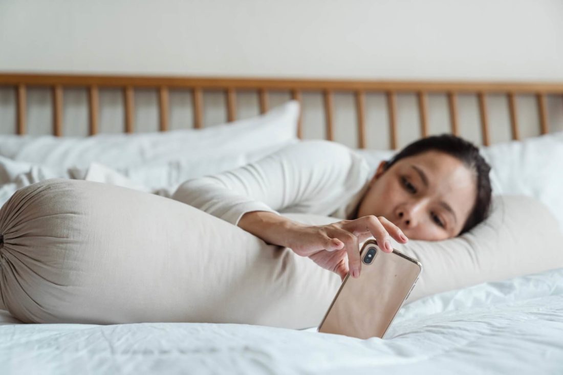 A woman in bed with a smartphone before long-distance movers near her move her belongings to another location 