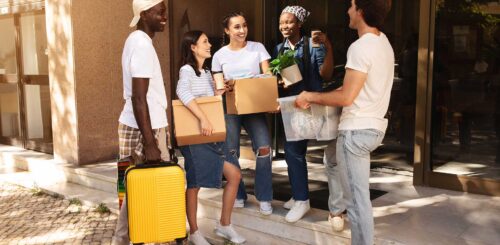 Happy multicultural group of young friends two guys and three ladies holding boxes with belongings and suitcase, standing by building, have conversation, student moving to new apartment, relocation